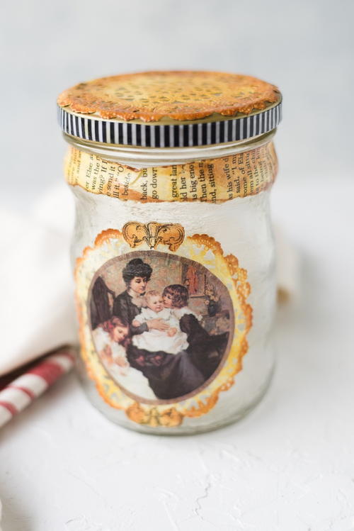 Multiple materials including a delicate paper doily paper cutouts fabric trim book page grunged with archival dye ink pads are used to decoupage a recycled glass jar with a metal lid The Ultimate Guide for How to Decoupage