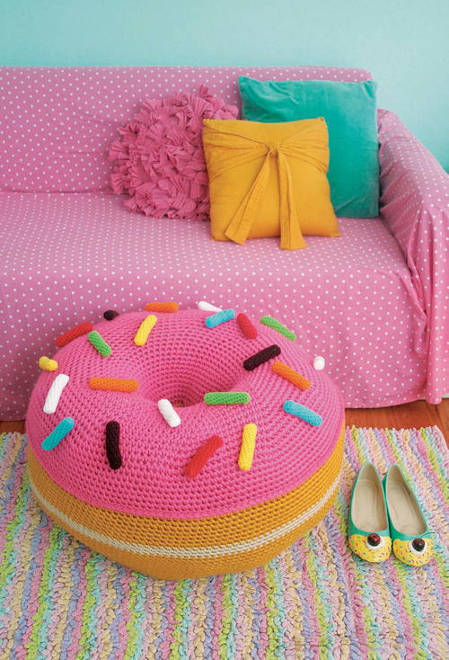 A large donut ottoman with pink frosting and colorful sprinkles sits in front of a couch covered in a pink polka dot slip cover. The donut pouf sits on a rainbow stripped rug.