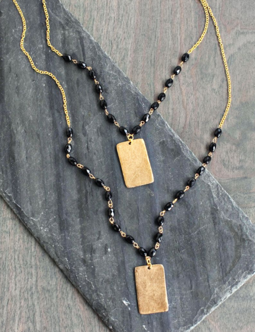 Two gold necklaces with black beads and a gold rectangular pendant laying on a grey stone slate. 